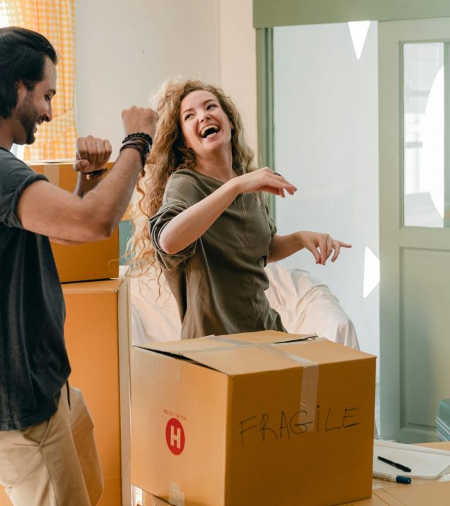 Cheerful laughing couple in casual clothes having fun and dancing together while unpacking carton boxes after moving into new contemporary apartment